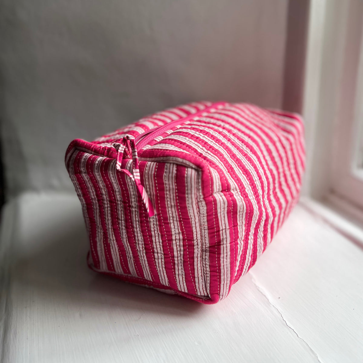 Harlequin quilted cotton washbag in pink stripe on a windowsill