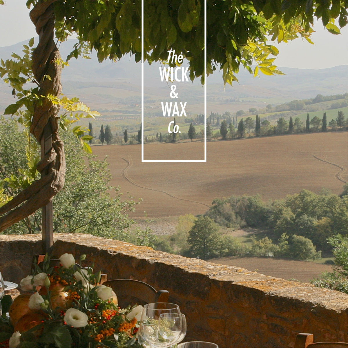 The Wick and Wax Co logo over a Tuscan countryside view